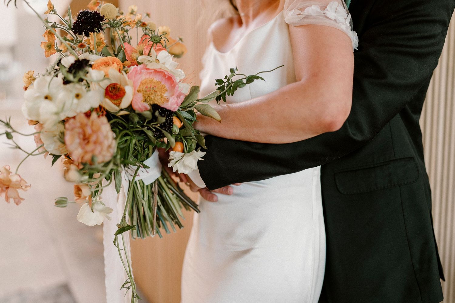 A wedding couple embraces while holding a lush bouquet of pink and white flowers.