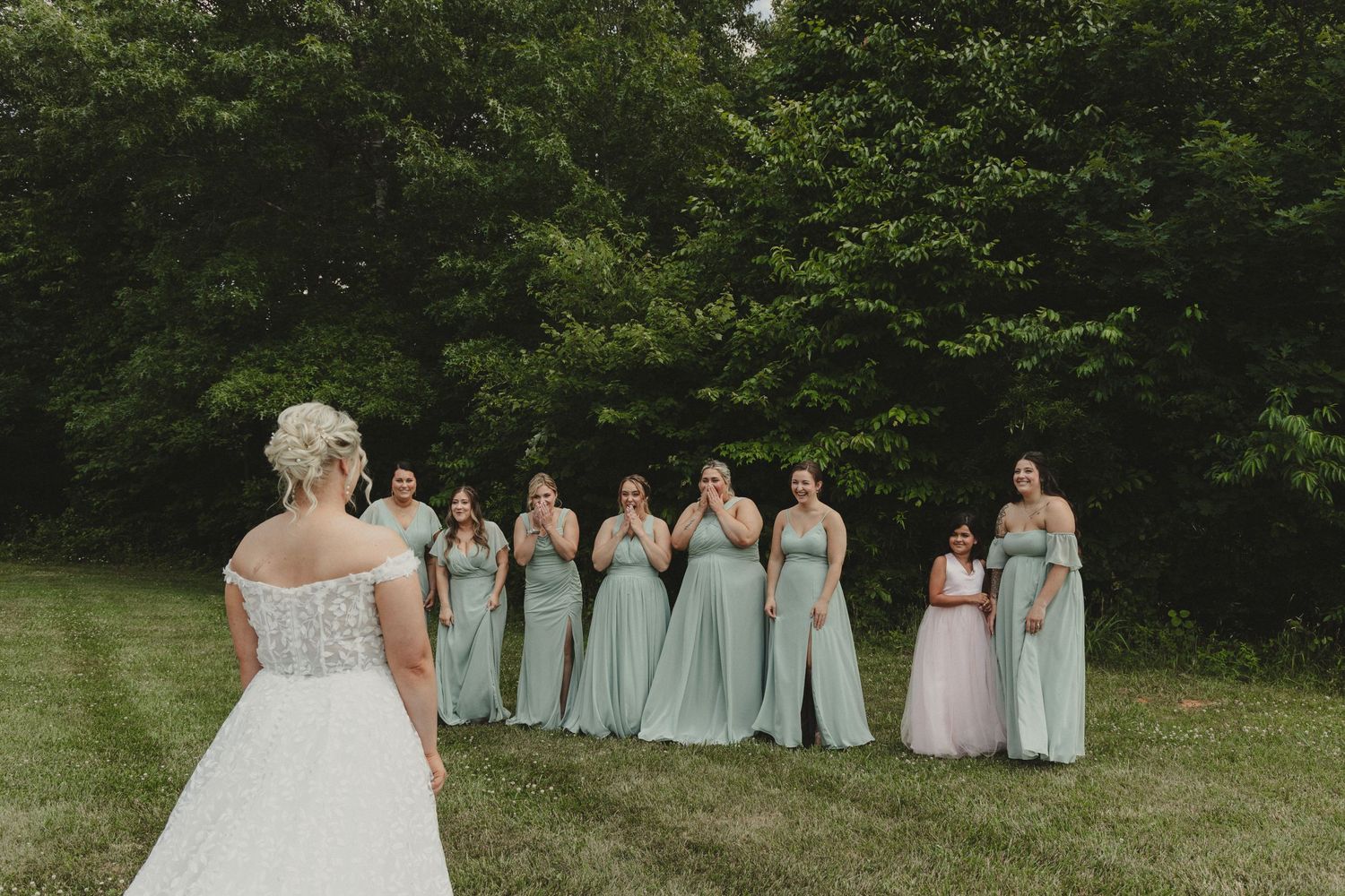 Bridesmaids in mint green dresses stand together on a lawn viewing the bride's dress.