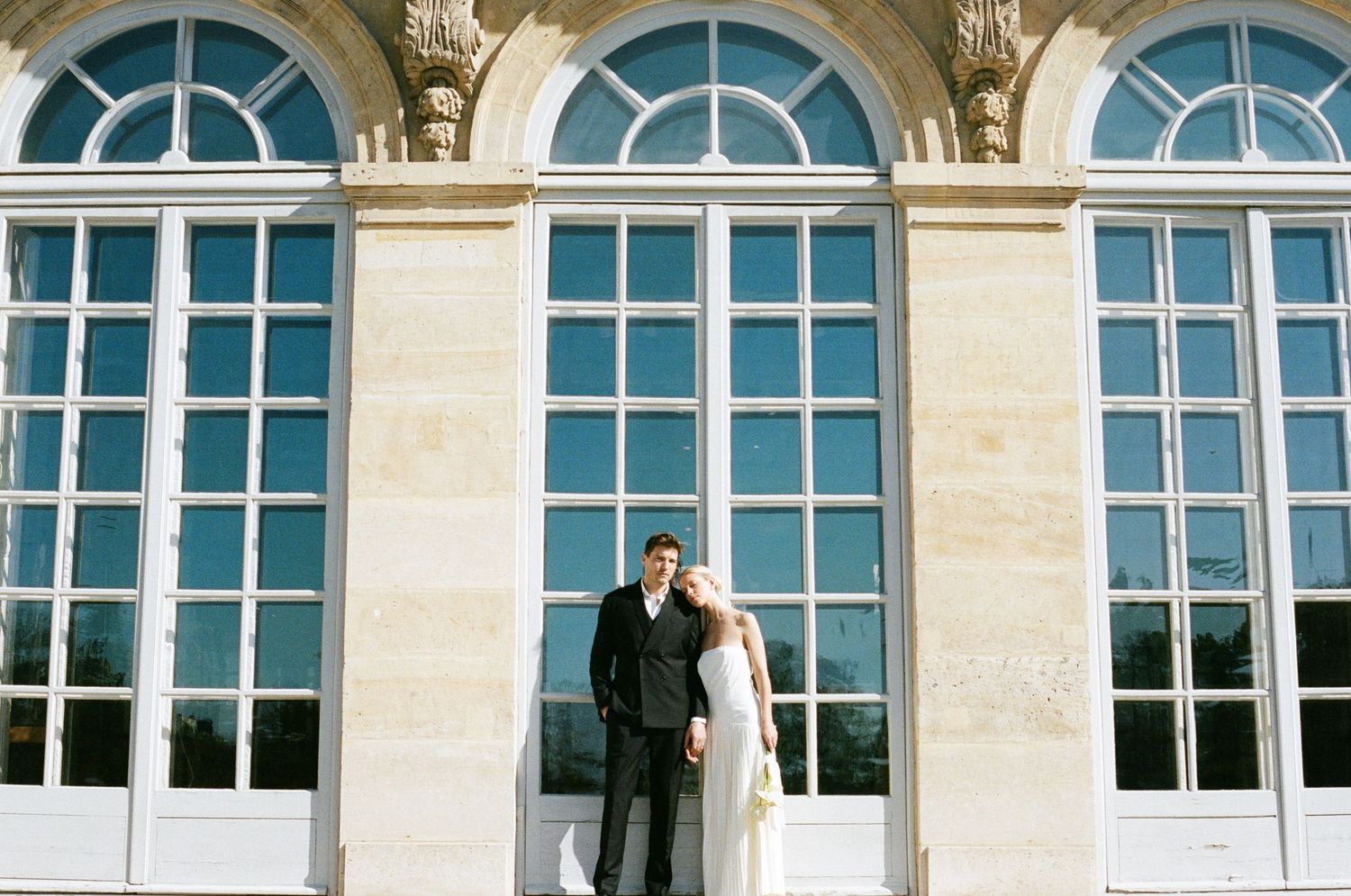 A couple in wedding attire stands in front of tall arched windows on a classic stone building facade.