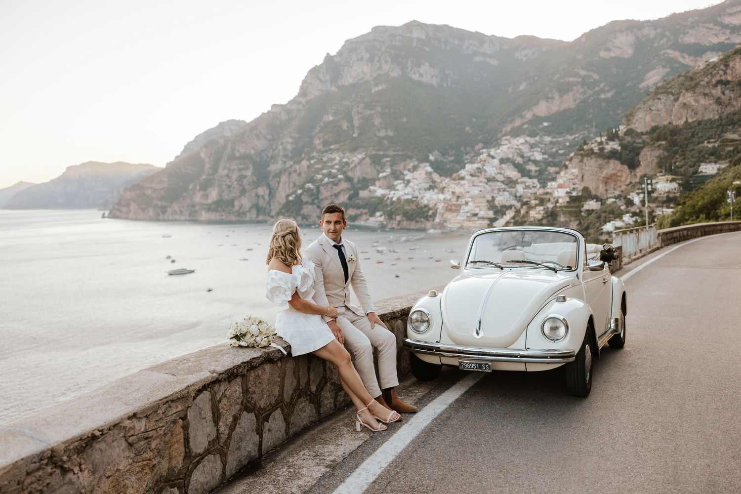 couple sitting on a stone wall next to a vintage fiat on their elopement day in Positano, Amalfi Coast, Italy
