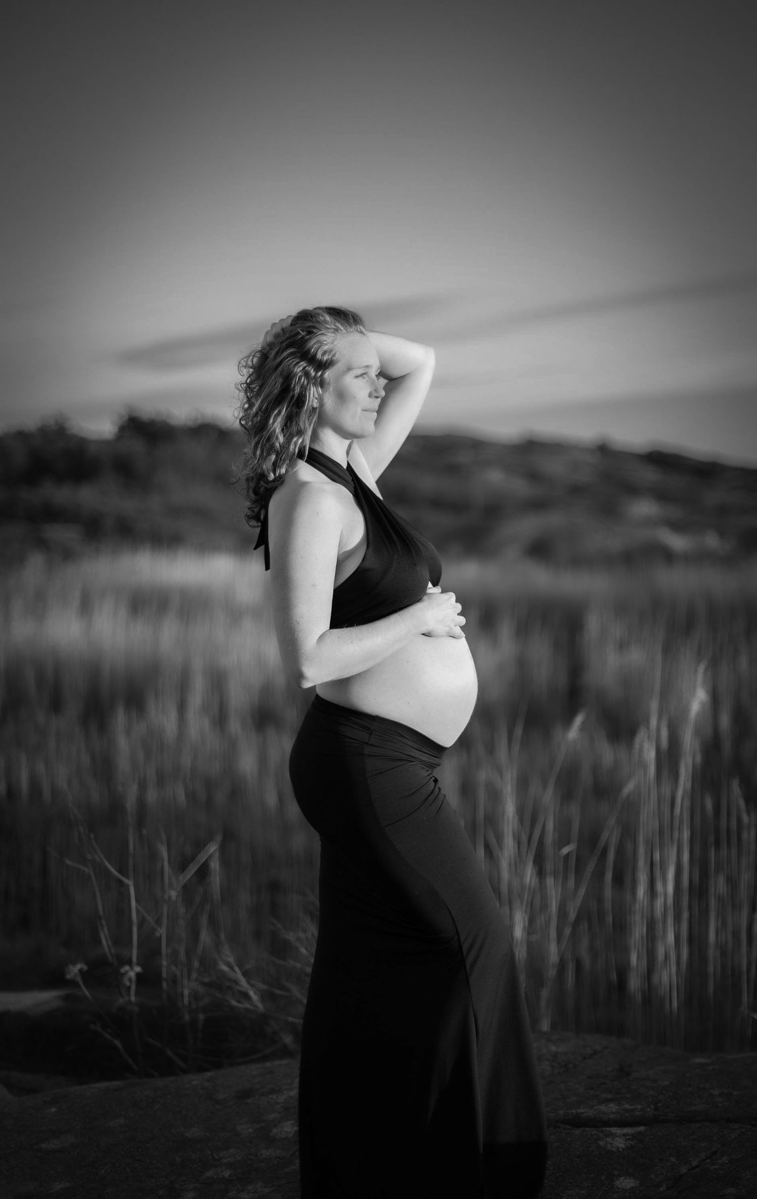 bild i svartvitt på en gravid kvinna som står och kollar utåt. hon har en svart topp och kjol på sig och håller på sin gravidmage.