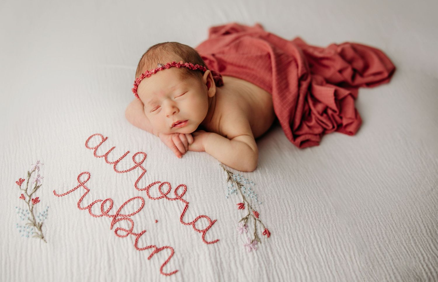 Sleeping newborn posed with red fabric and delicate embroidered text on white background.
