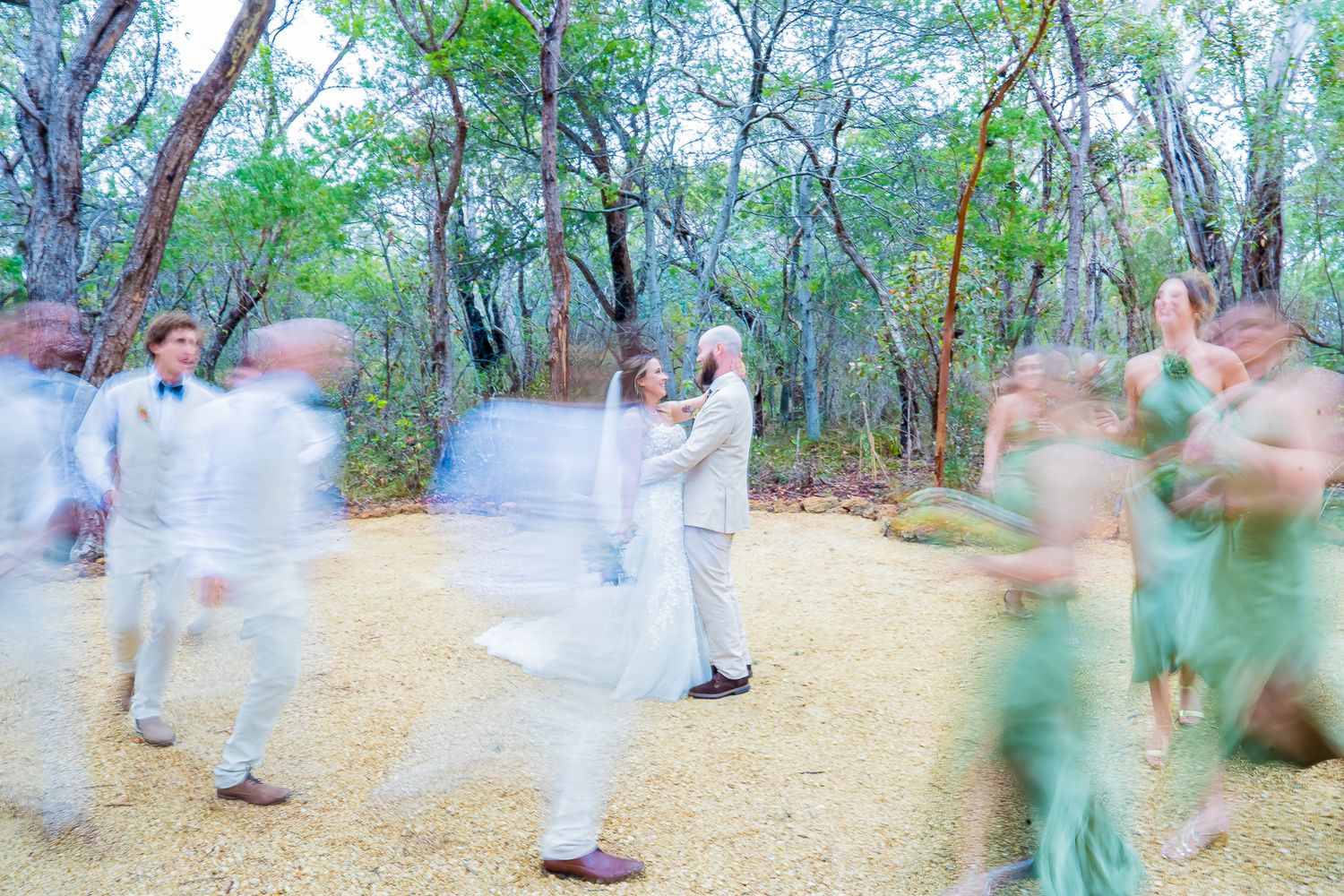 Long exposure photograph showing blurred motion of dancers in white and green clothing moving on a dirt path in a wooded area.