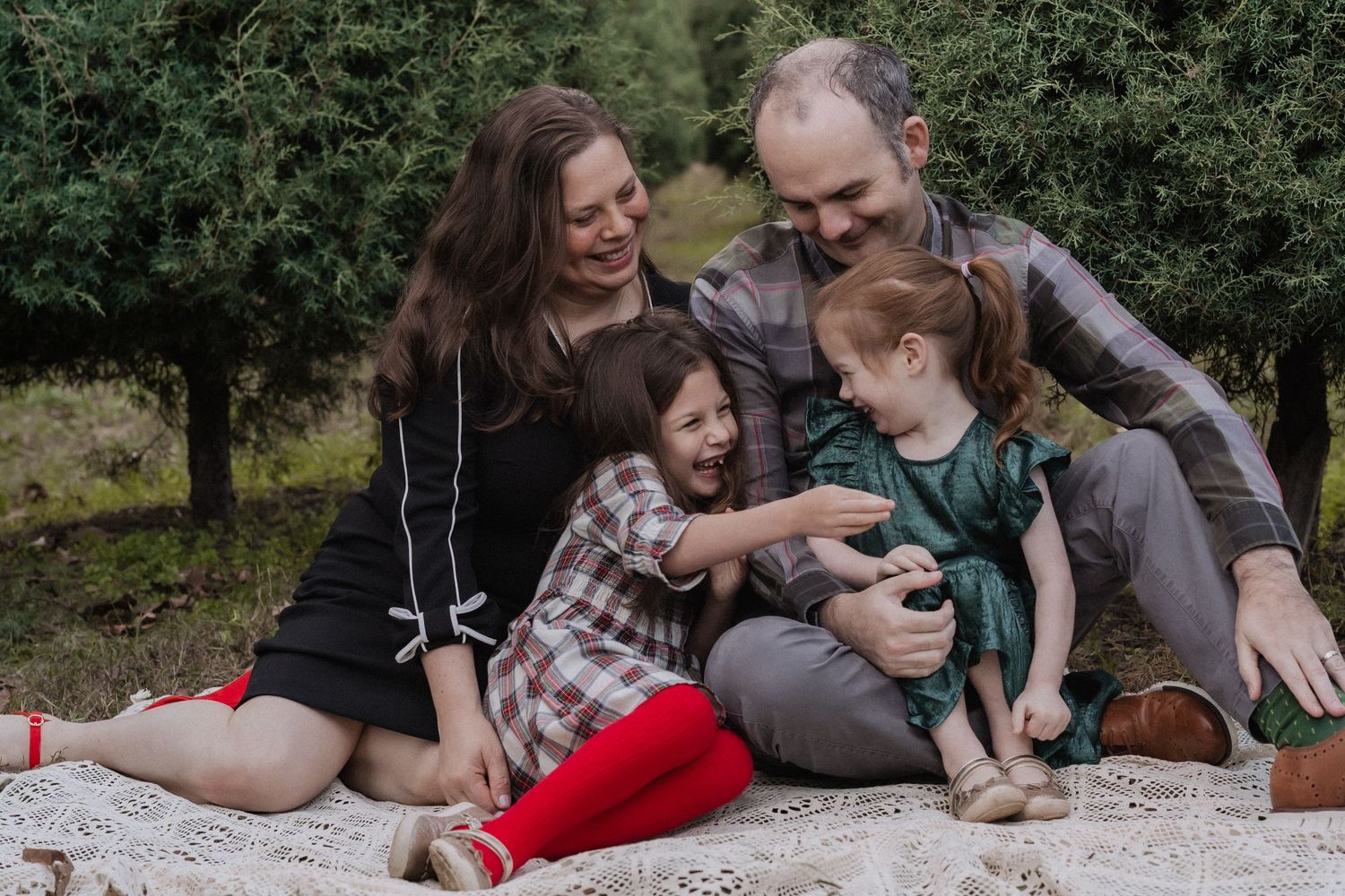 A family sits together on a blanket outdoors having a candid moment among trees and greenery.