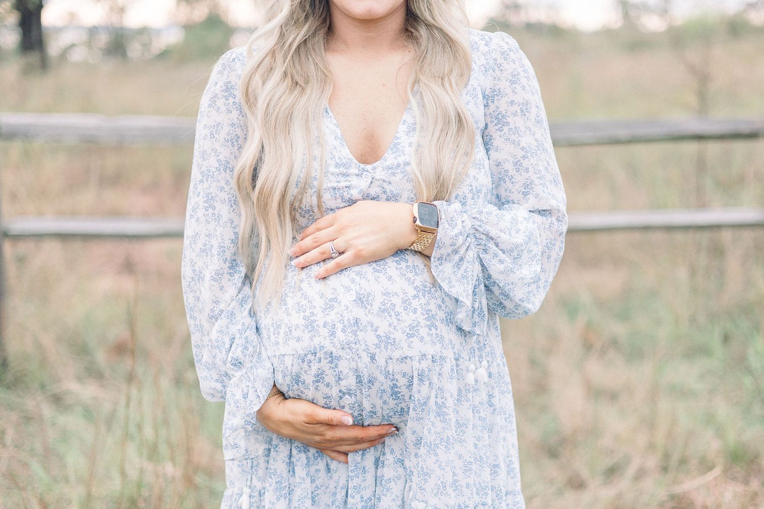 Soft blue floral maternity dress with bell sleeves shown in outdoor field setting with wooden fence. Aiken Maternity Photographer.
