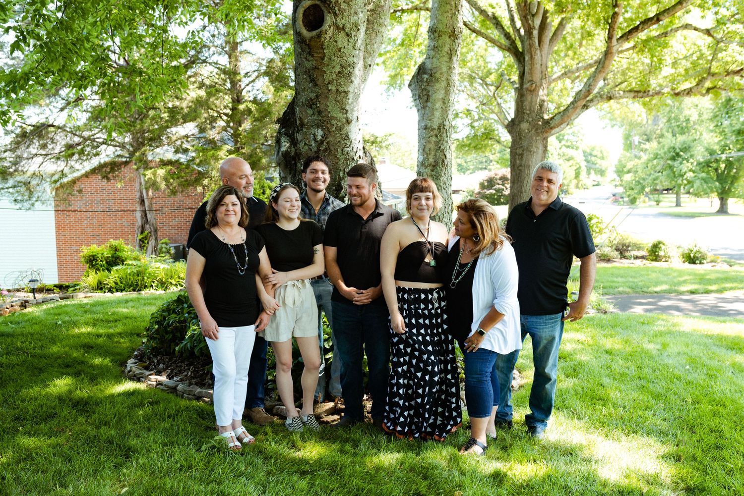 Group photo taken under large tree with sunlight filtering through branches on grassy lawn.