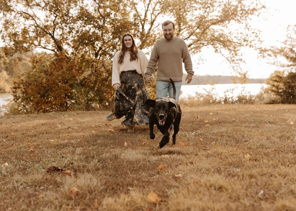 A couple walking their black dog on an autumn path near a lake during golden hour with warm sepia tones.