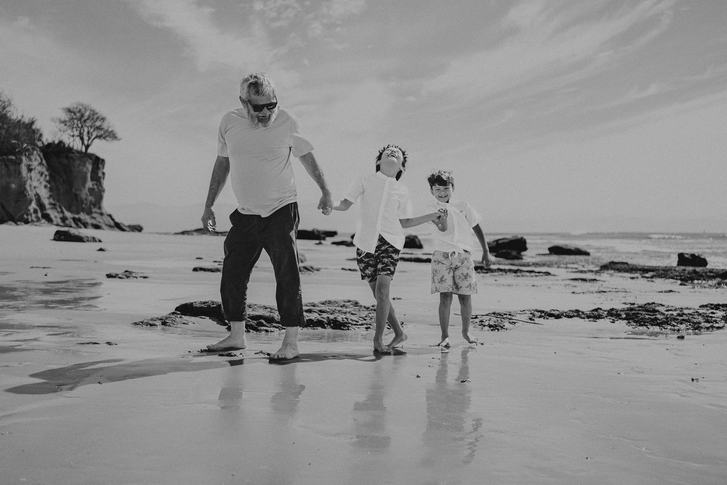 Three people holding hands while walking on a rocky beach in black and white.