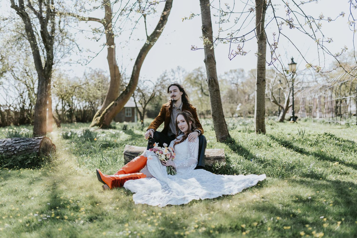 A figure in a black jacket and flowing white dress sits on a blanket in a sunlit spring forest meadow.