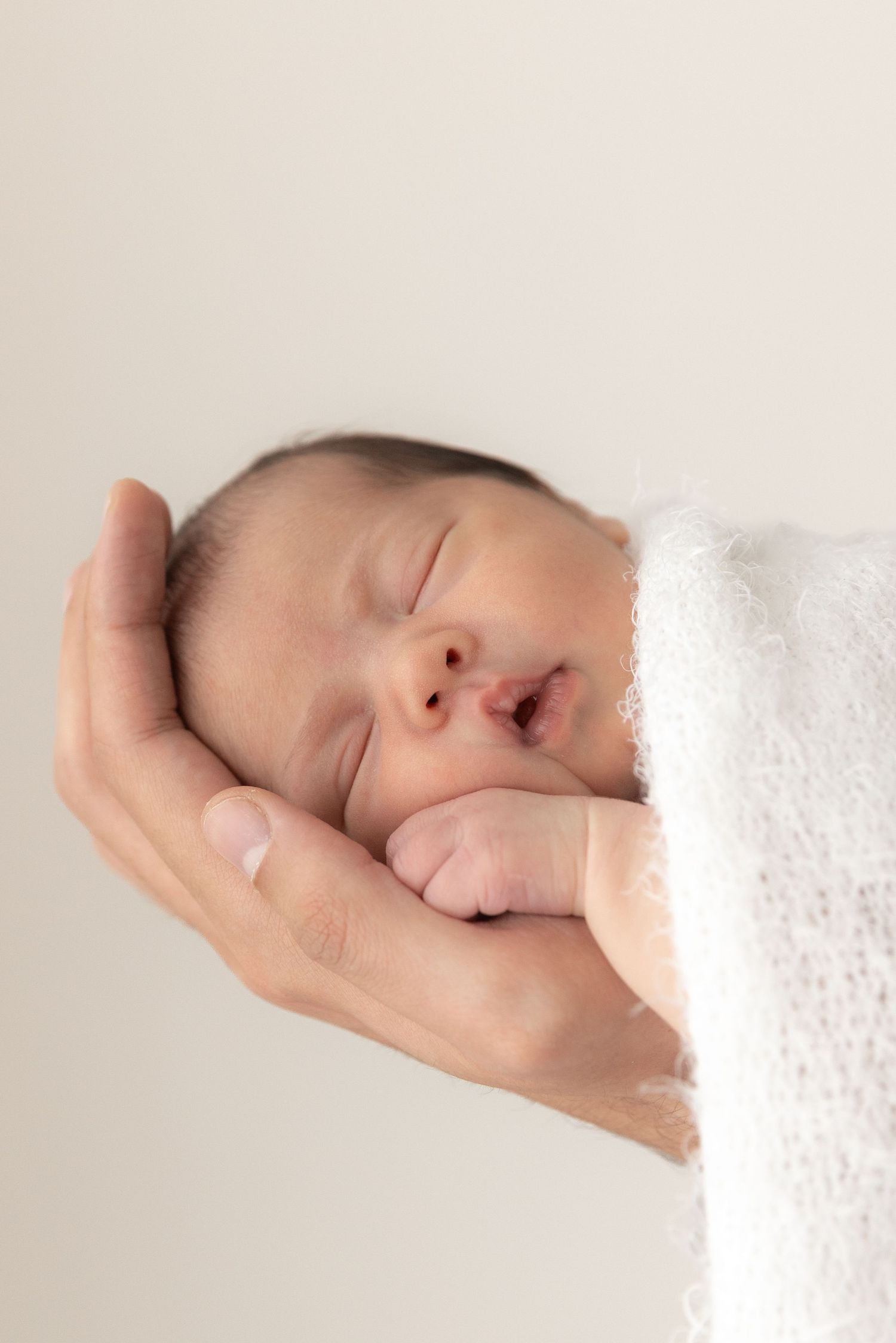 Sleeping newborn baby wrapped in white blanket rests peacefully in adult's hand against neutral background.
