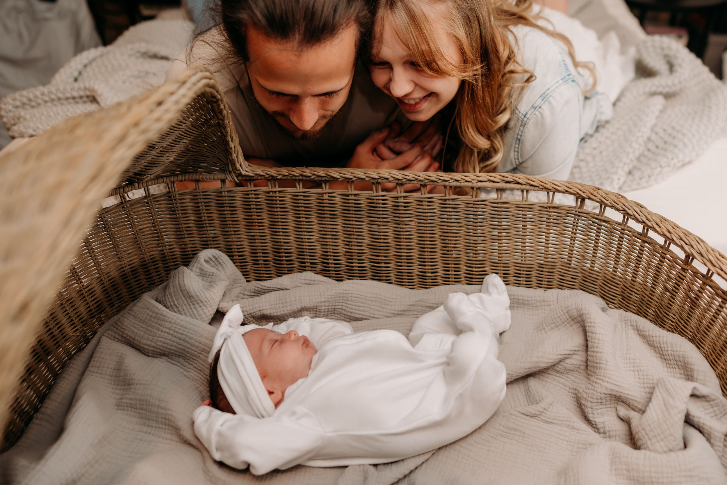 Newborn baby sleeping peacefully in wicker bassinet while parents lovingly look on from above during in home newborn photos.