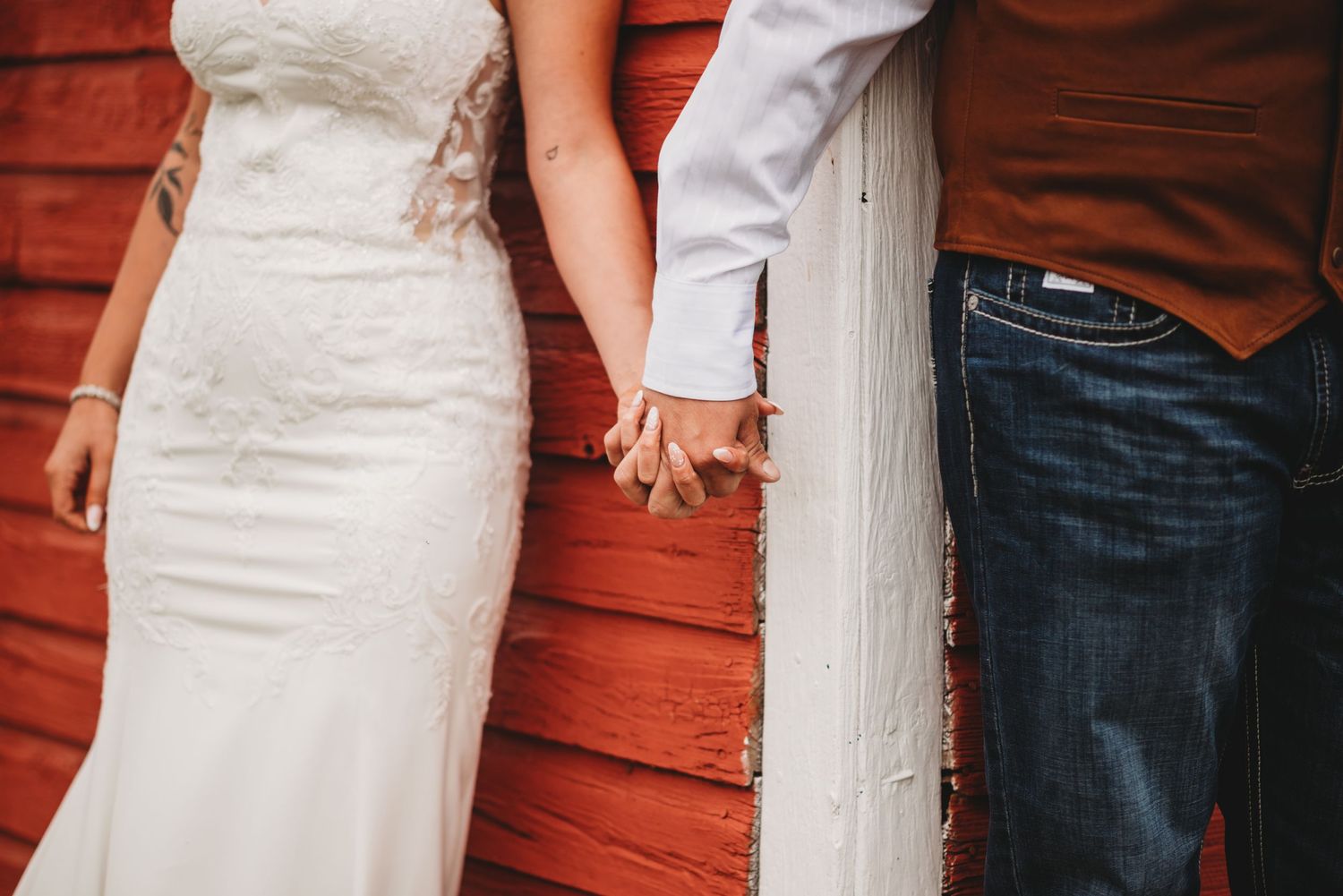 Close up of held hands against a rustic red barn wall with wedding attire visible.