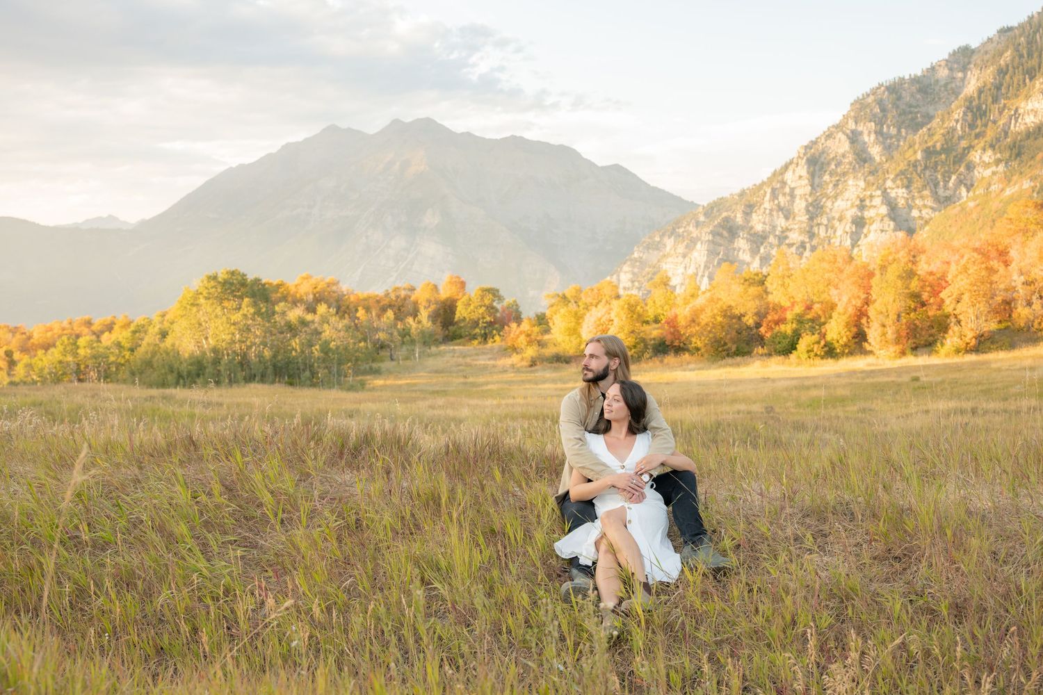 A couple embraces in a golden meadow with majestic mountains and autumn foliage in the background at sunset.
