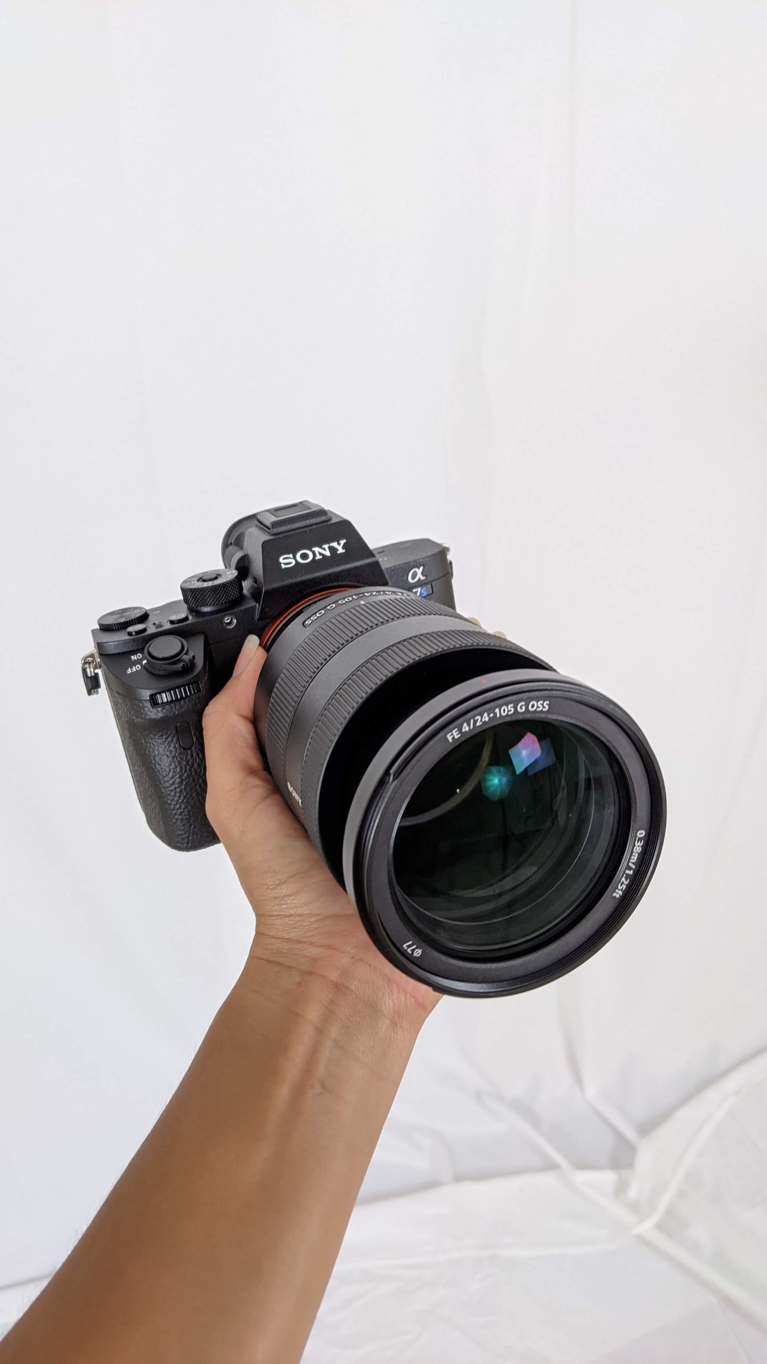 Hand holding a Sony Alpha mirrorless camera with a 24-105mm G OSS lens against a white background.