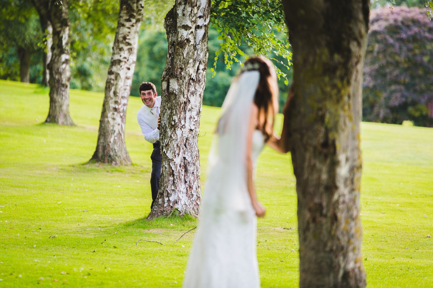 A bride in a white wedding dress walks through a lush green garden setting with tall trees and pink flowers.