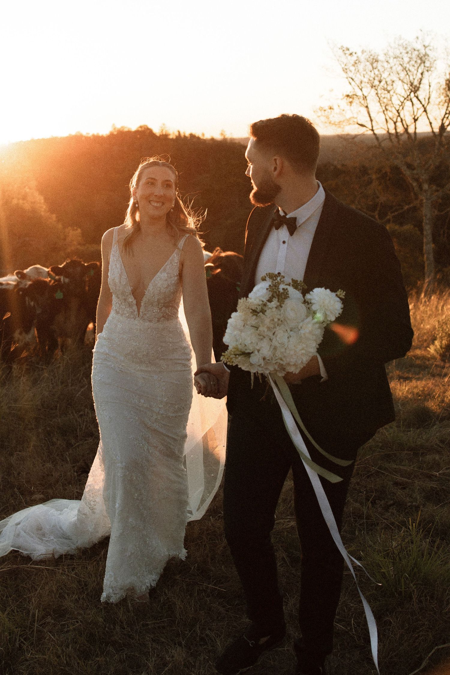 A bride and groom share a romantic moment at sunset in a rustic outdoor setting with white floral bouquet.