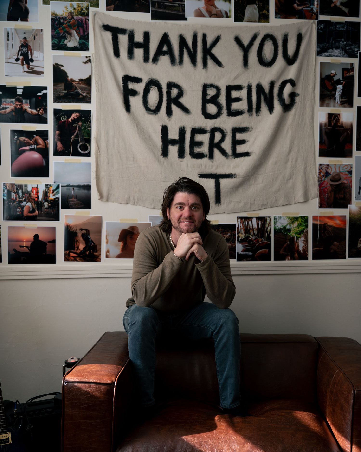 A cozy room with a white banner reading 'THANK YOU FOR BEING HERE' hanging above a brown leather couch with photos on the wall.