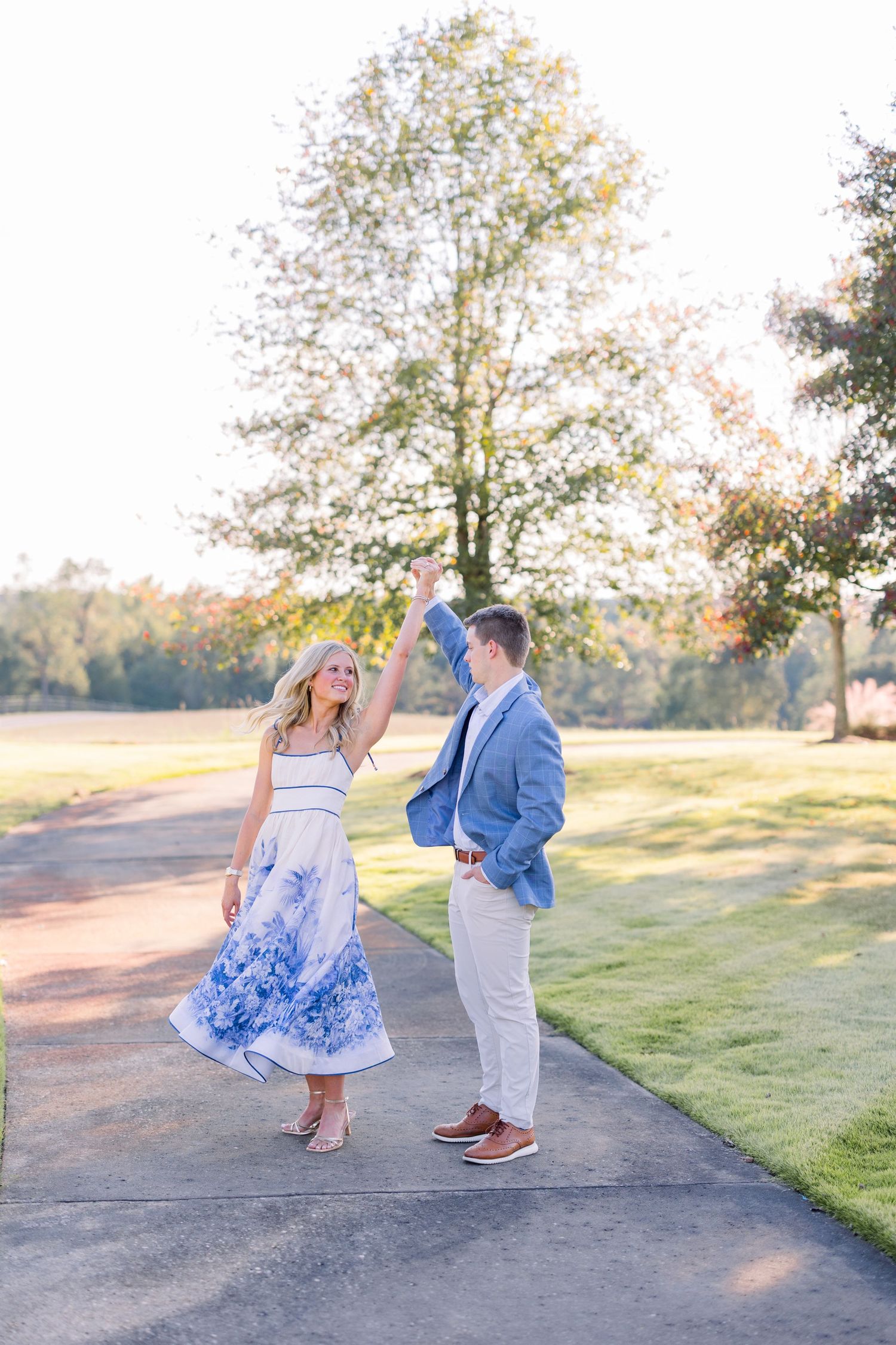 A couple in blue and white attire dances playfully together on a sunlit path surrounded by green grass and trees.