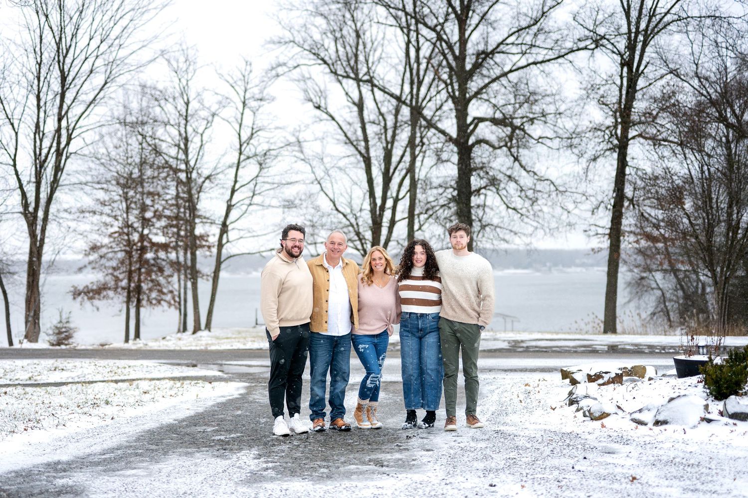 Happy family of six posing together outdoors in winter snow near a frozen lake with bare trees.