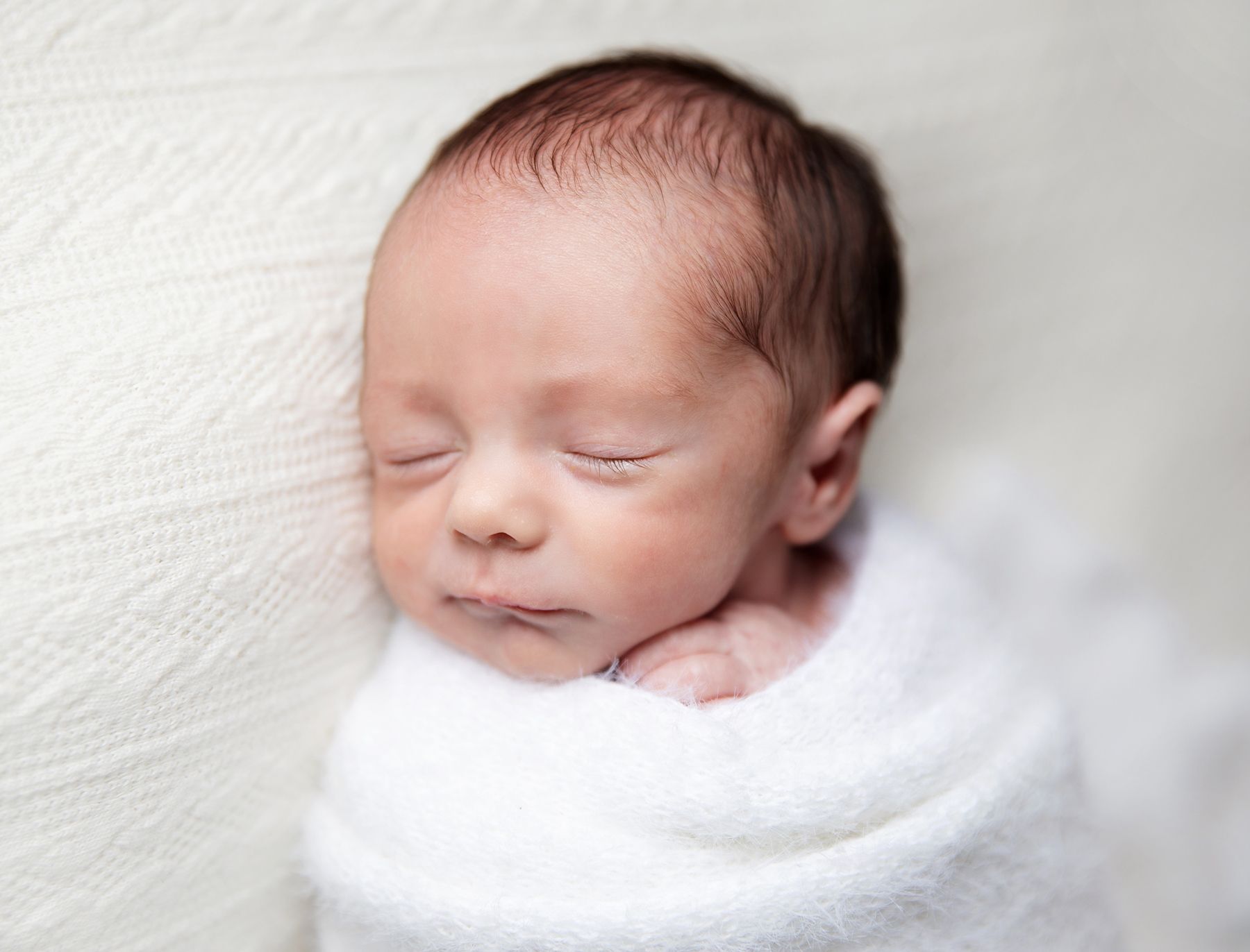 Peaceful newborn sleeps while wrapped snugly in soft white blankets creating a serene portrait. During a newborn session in Bexley