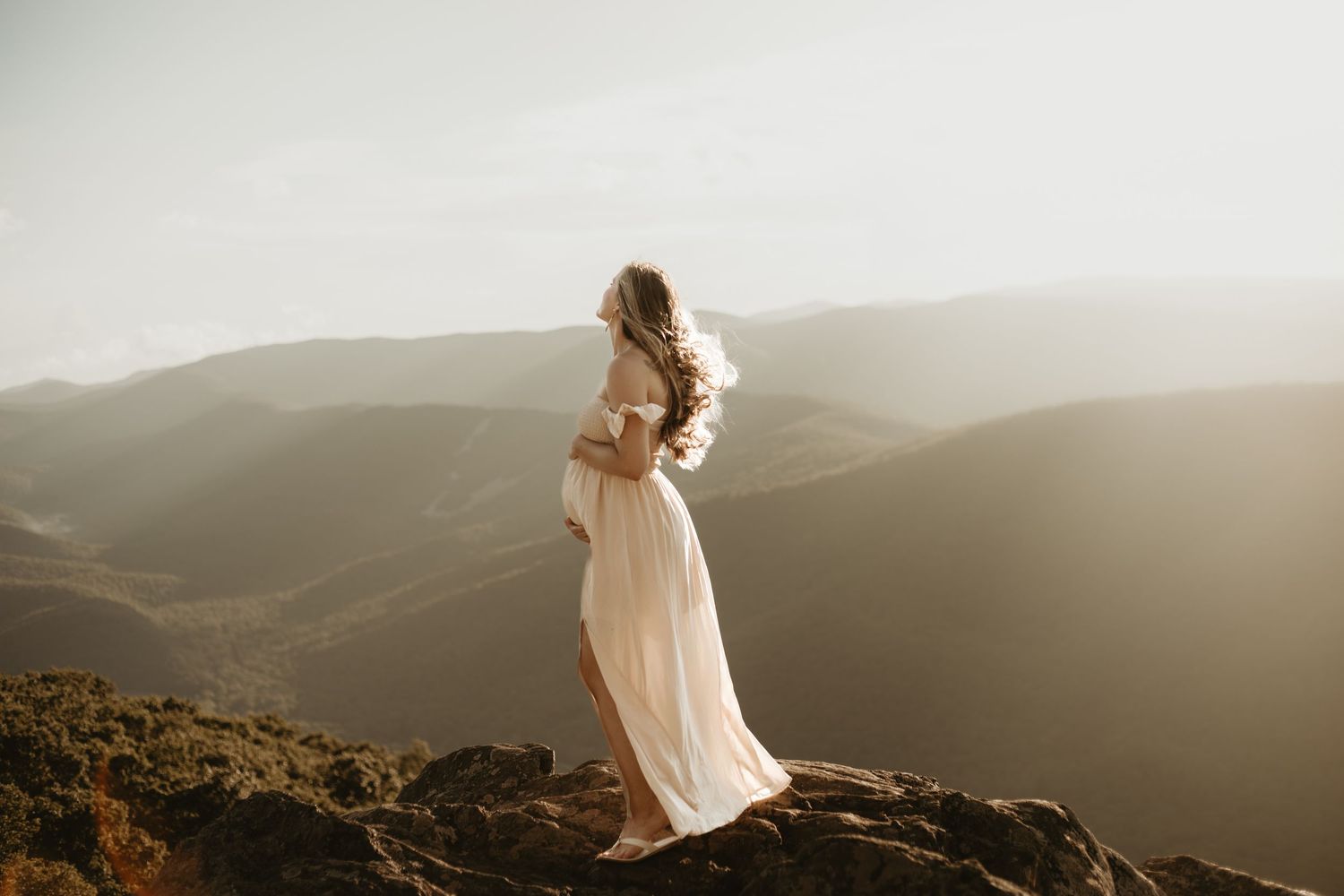 A figure in a flowing white dress stands atop a mountain ridge at sunset, silhouetted against misty rolling hills.