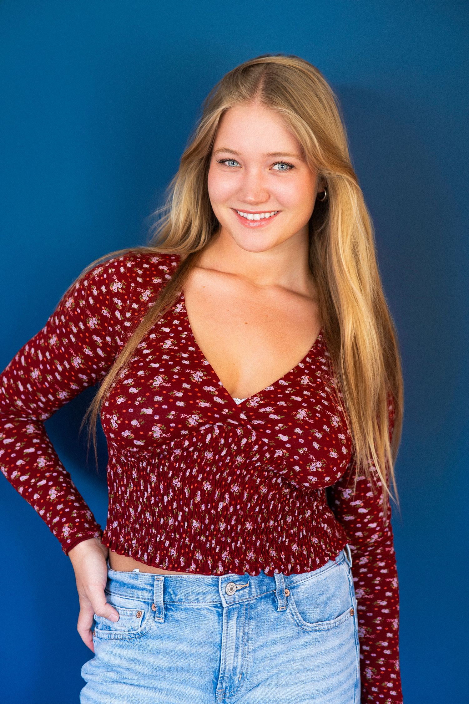 Young woman in burgundy floral print v-neck top and light blue jeans poses against bright blue background.