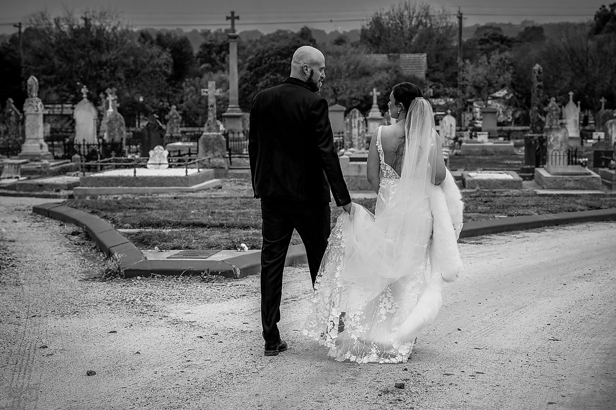 A black and white photograph of a couple walking down a path in a cemetery on their wedding day.
