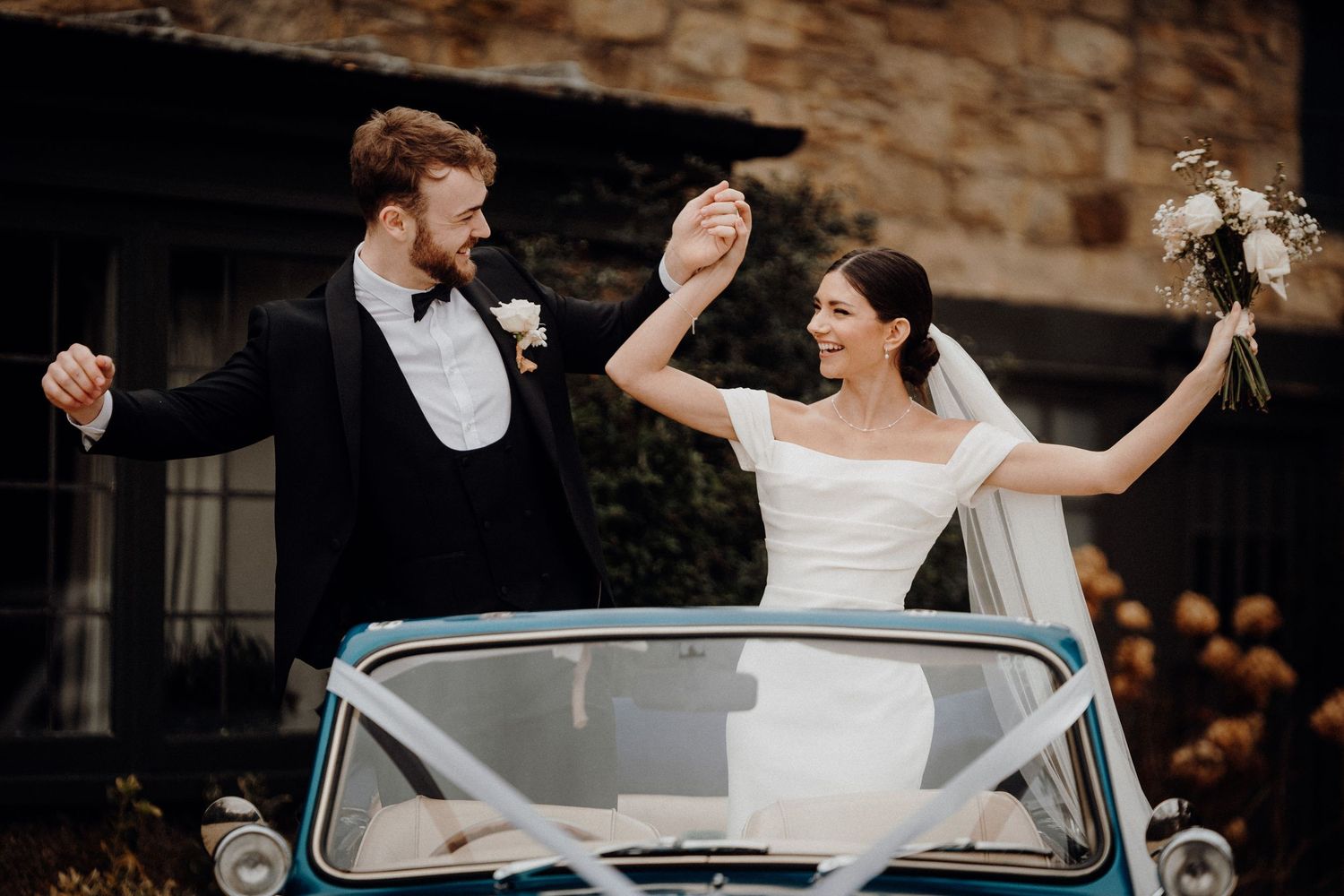 A couple celebrates their wedding day with joyful dance moves in front of a stone building.