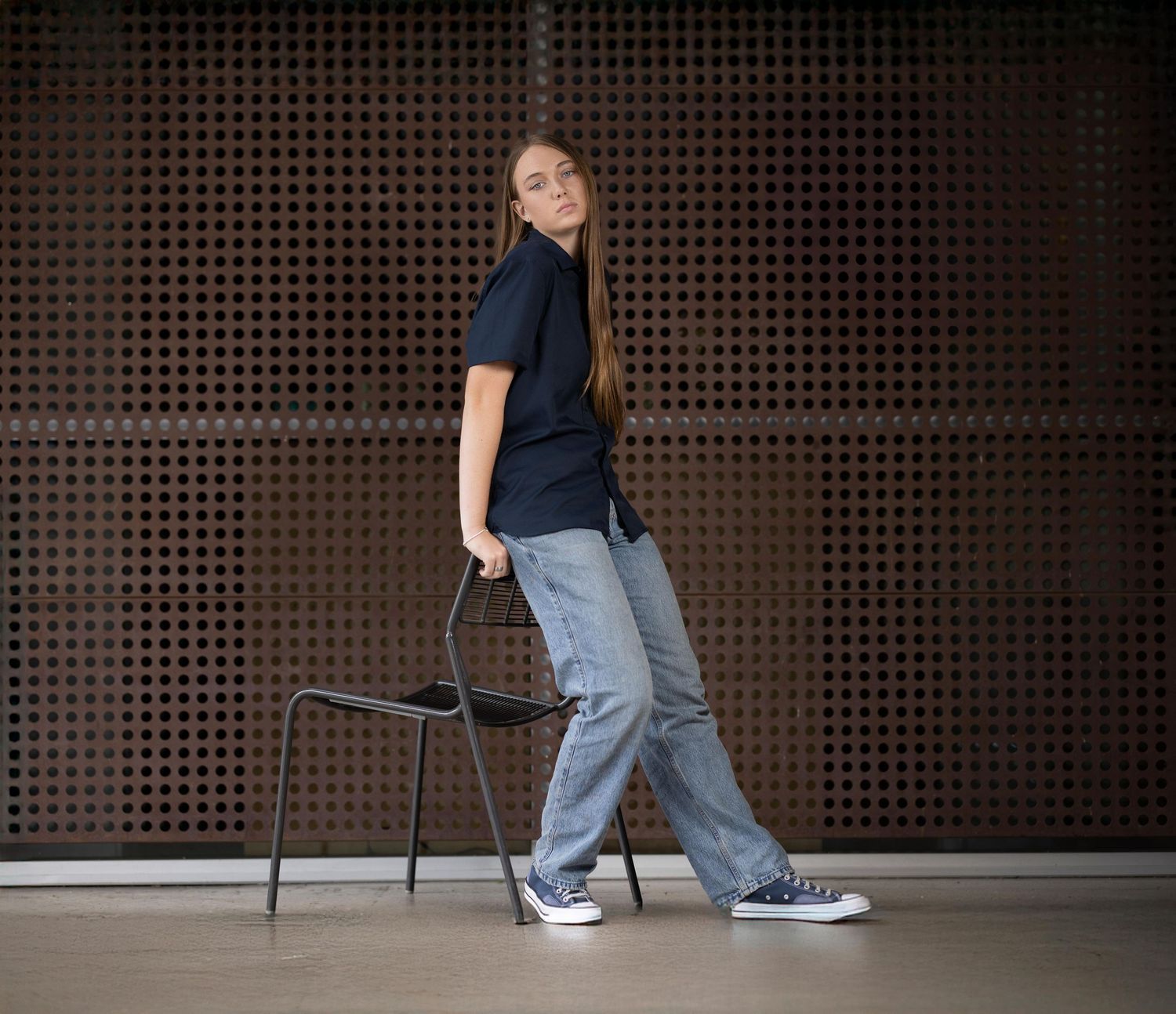A young woman in blue shirt and jeans pose against a dark perforated wall with chair casting dramatic shadows.