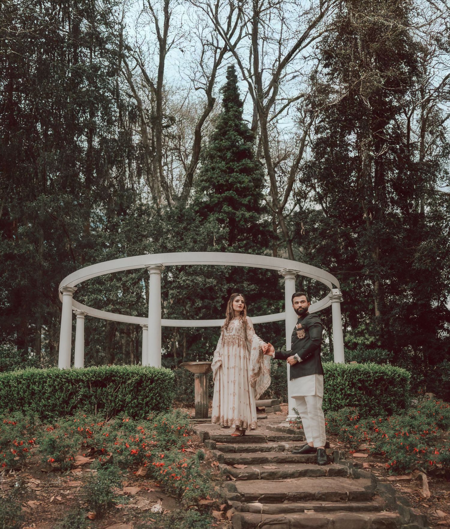 Two people stand on rustic stone steps surrounded by lush greenery and red flowering plants in a garden setting.