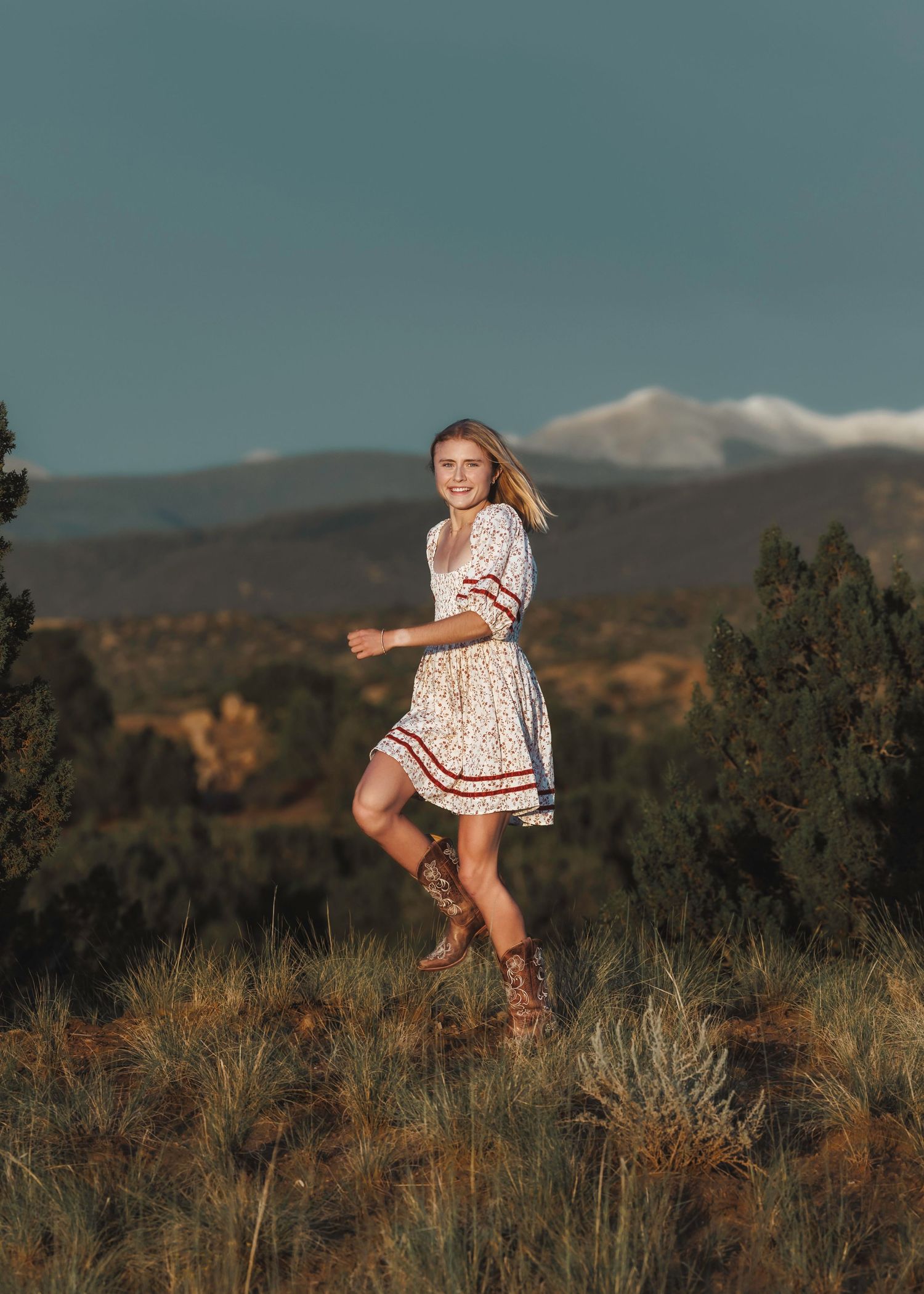 Santa Fe senior in floral dress and cowboy boots dancing in mountain landscape under blue sky.