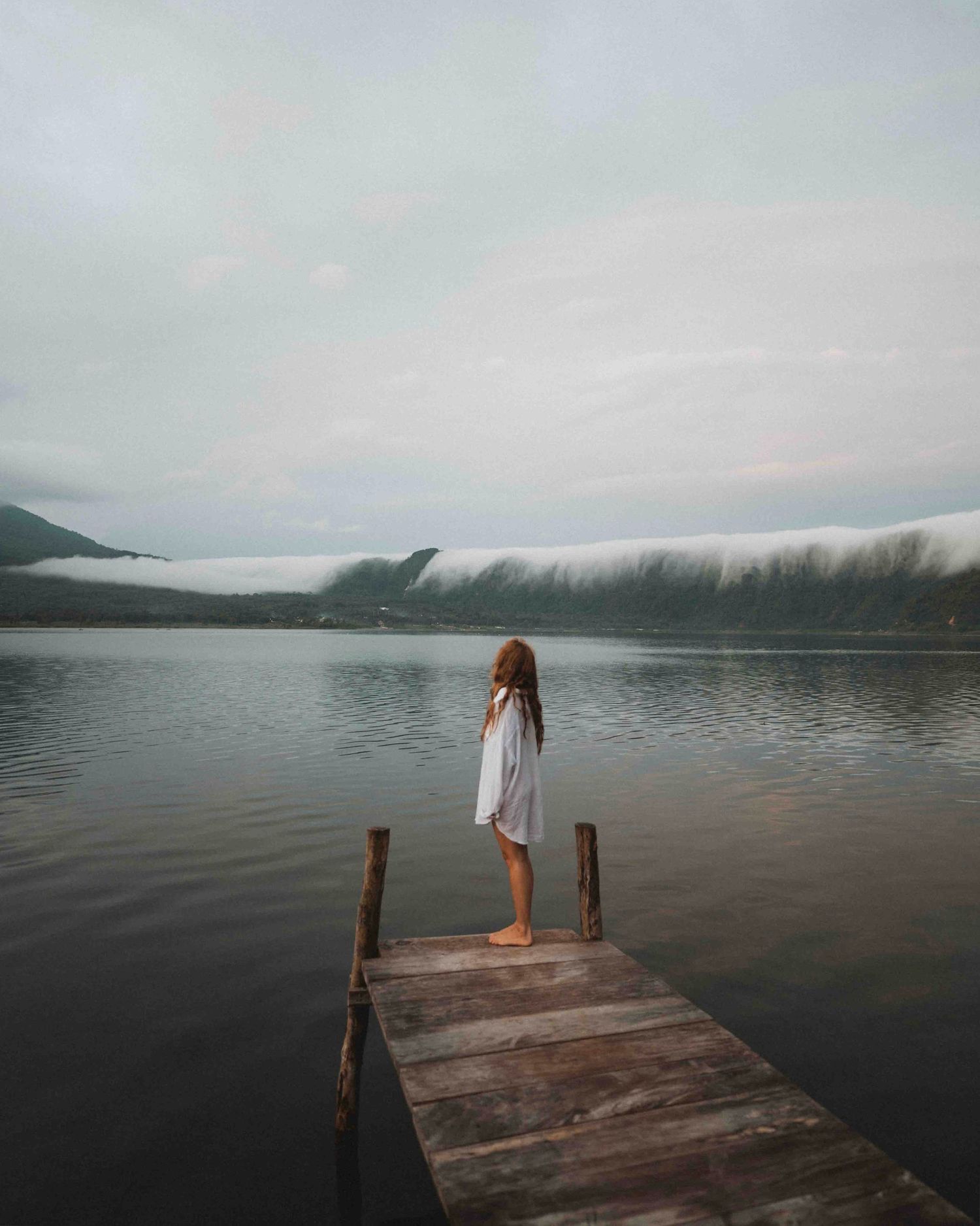 A wooden dock extends into a misty lake surrounded by mountains on an overcast day.