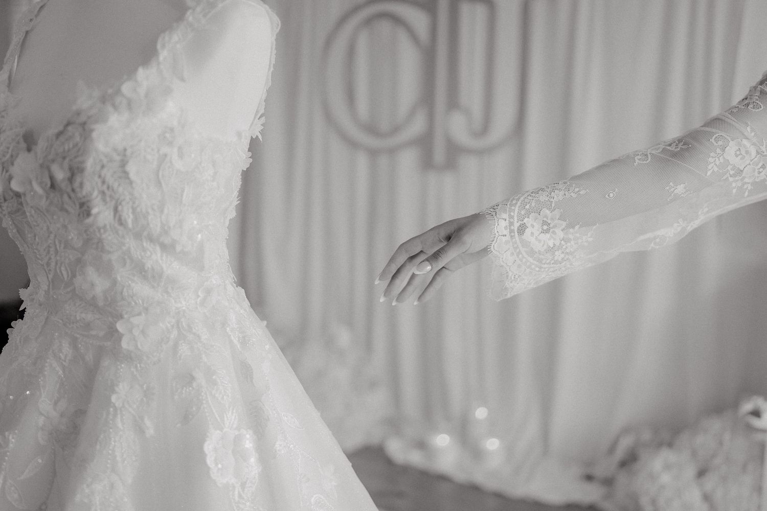 Black and white photo of a bride in a lace wedding dress standing in front of a window with soft lighting.