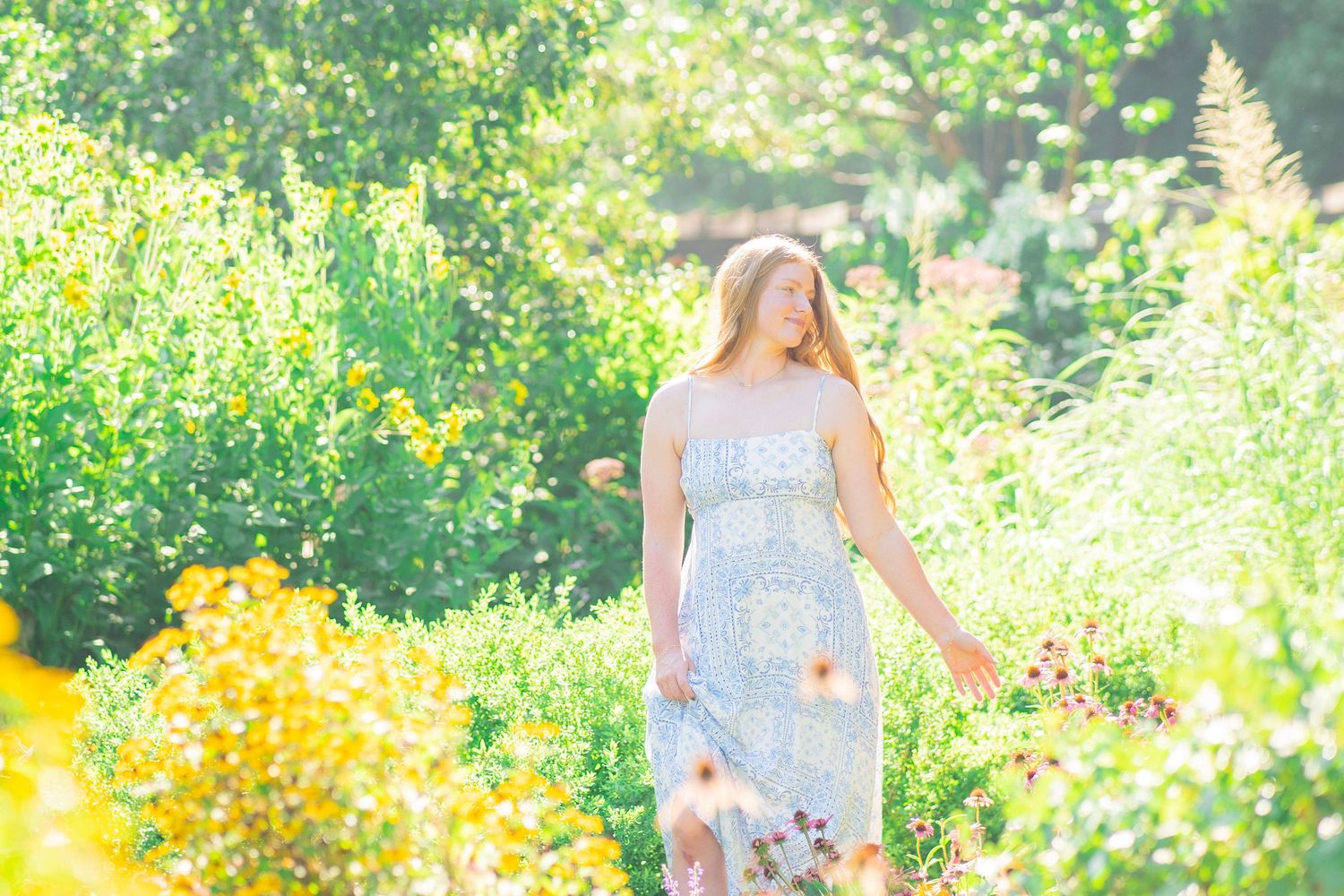 Young blonde woman in floral sundress walks through vibrant sunlit wildflower garden.