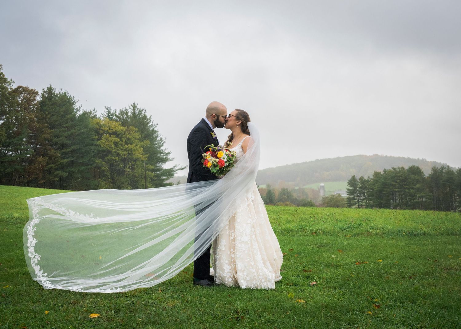 Newlyweds share an intimate moment outdoors with autumn foliage and rustic architecture in background.