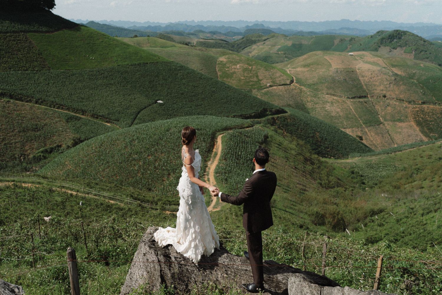 A couple in wedding attire stands on a mountaintop overlooking rolling green hills on their special day.