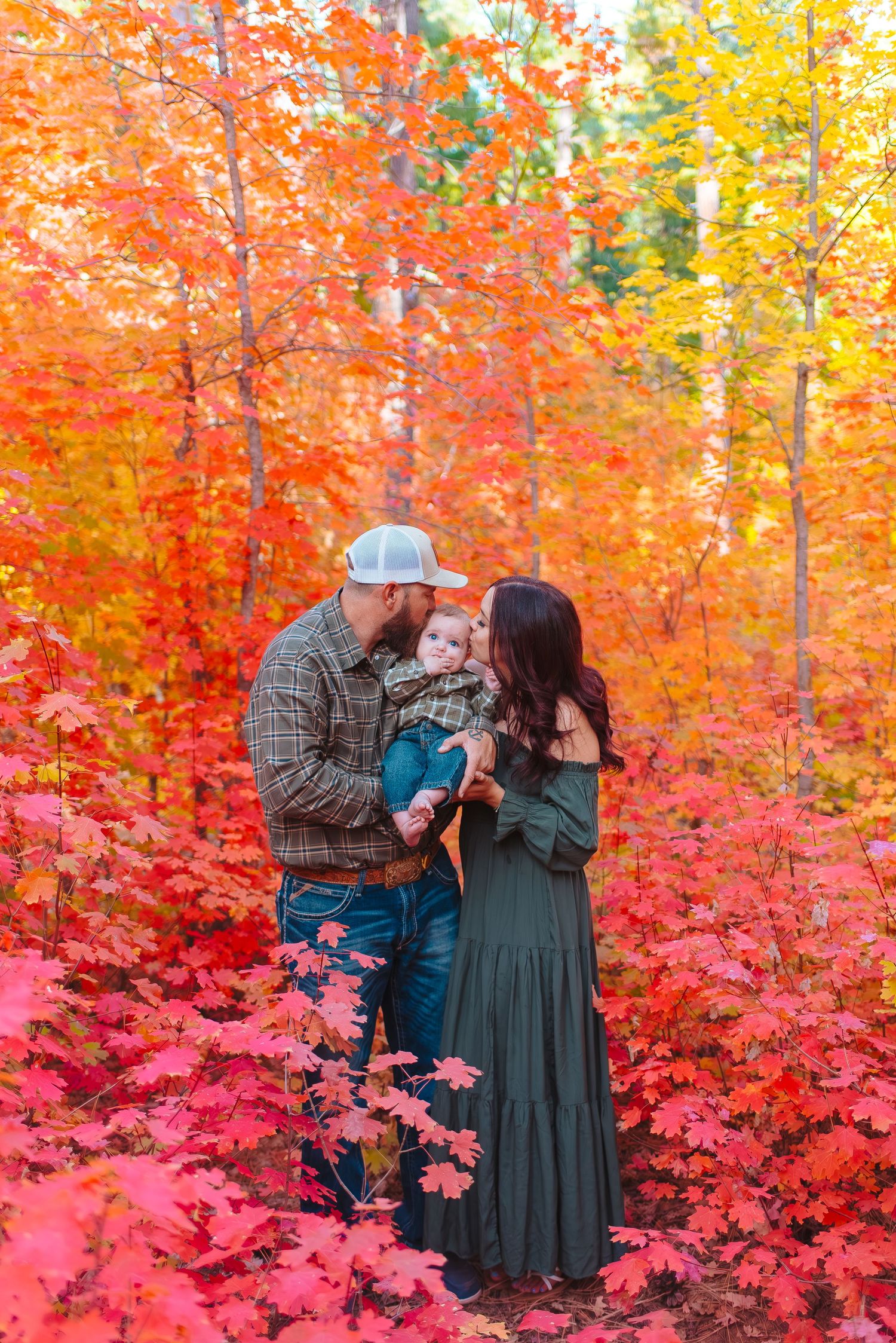 A family shares an intimate moment surrounded by vibrant red and orange fall foliage in a romantic forest setting.
