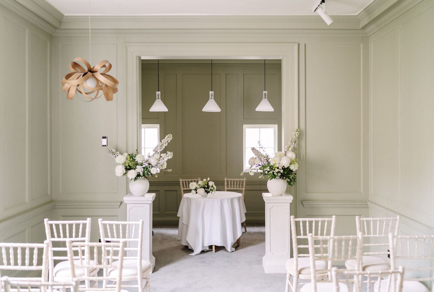 Elegant wedding ceremony space with white chiavari chairs, pendant lights, and floral arrangements on pedestals.
