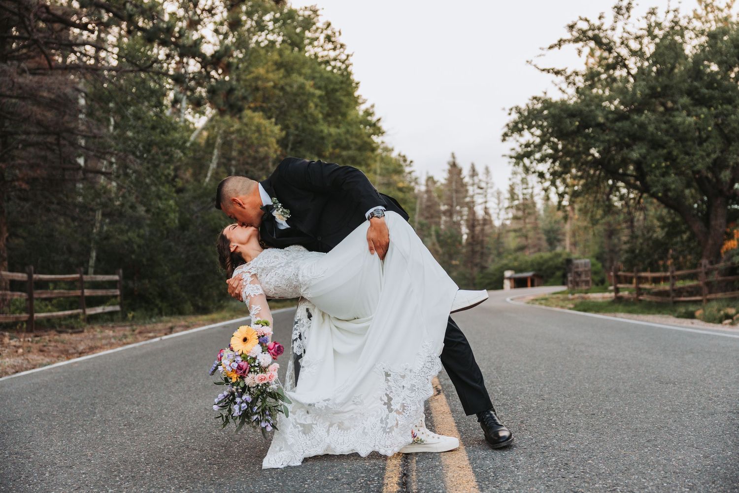A couple in wedding attire embraces while walking down a tree-lined road during sunset.
