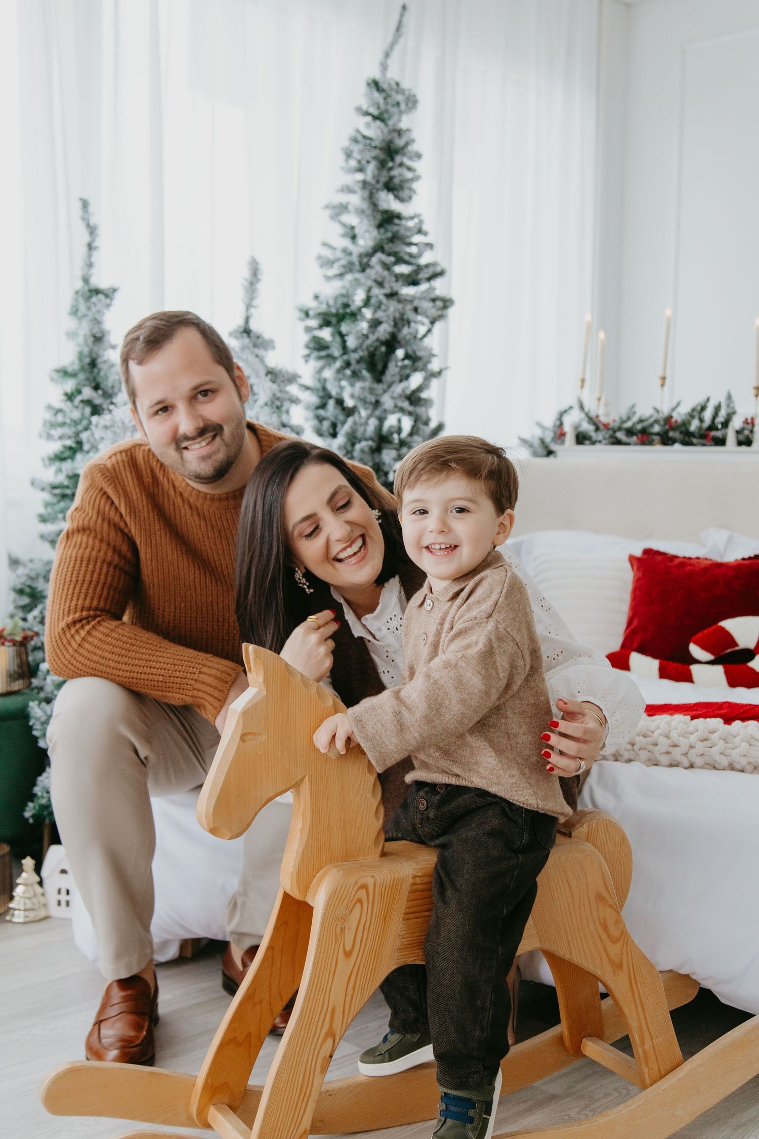 A family shares joyful moments around a wooden rocking horse during a holiday photoshoot with Christmas trees in background.