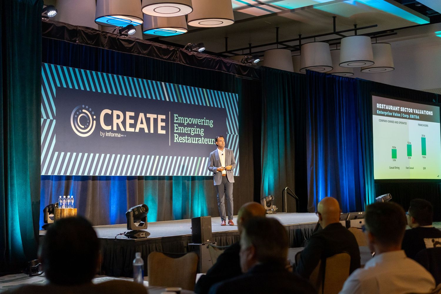 Speaker presents at CREATE Empowering Emerging Restaurateurs conference on a blue-lit stage before an engaged audience.