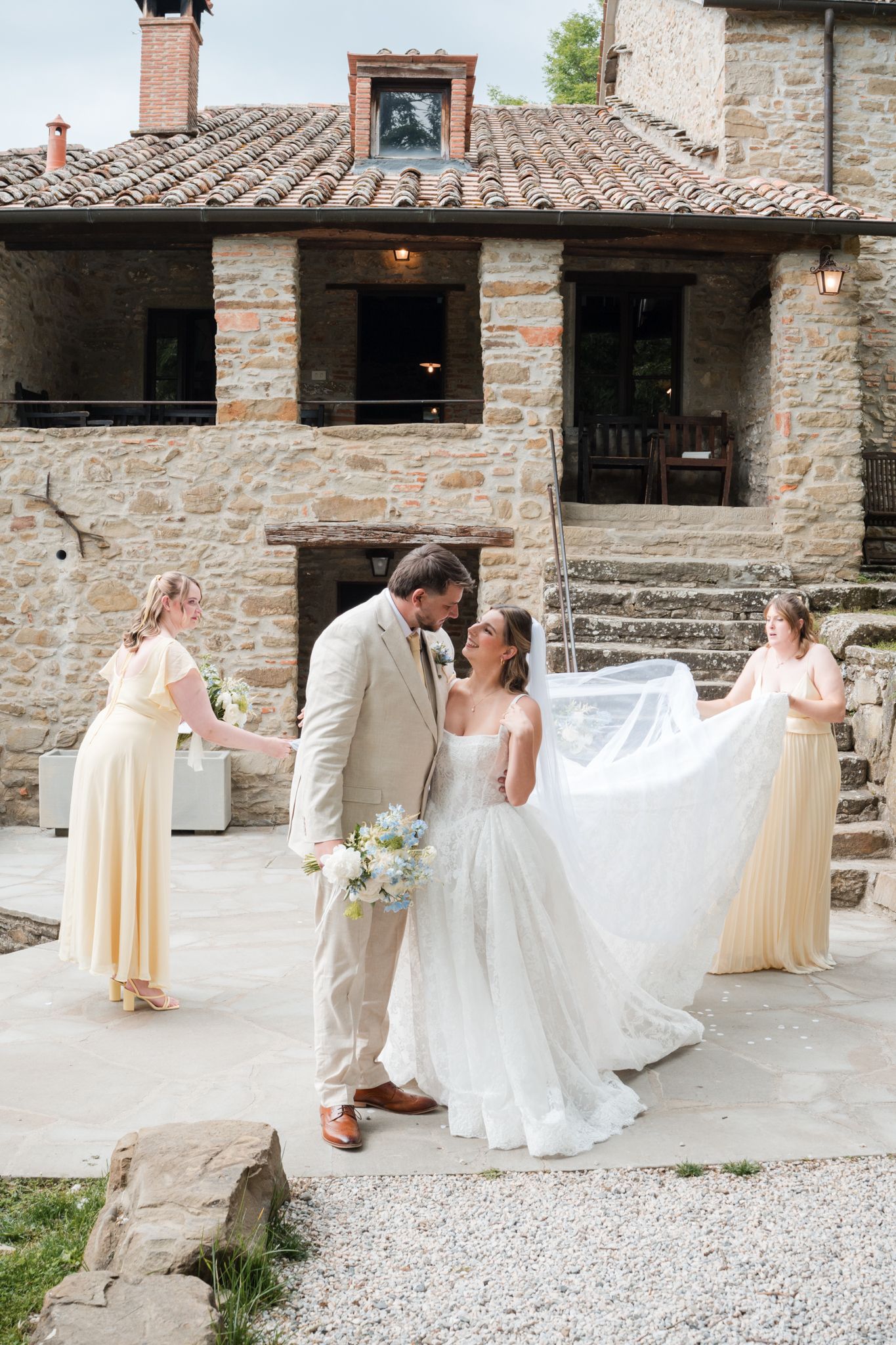 Bride and groom share a kiss outdoors at a rustic stone villa with bridesmaids in yellow dresses.