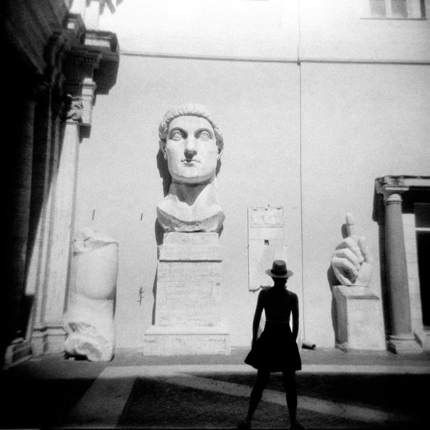 Black and white photo of a woman in a hat gazing at a giant Roman bust sculpture in a museum courtyard.