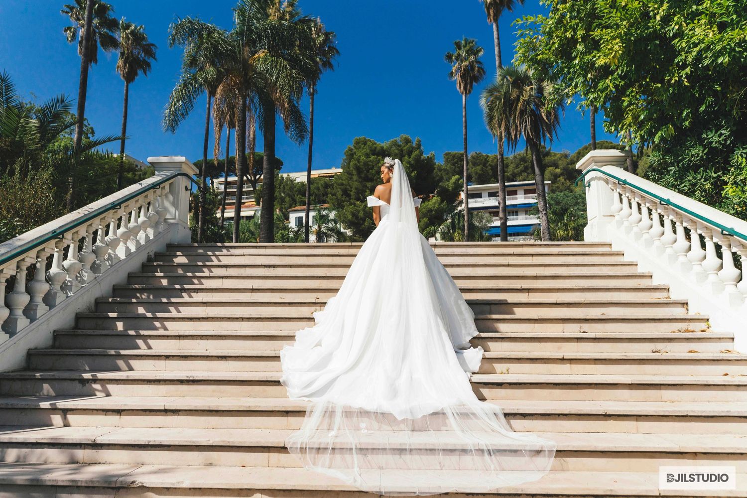 A flowing white wedding dress with a long train cascades down elegant marble stairs lined with palm trees under blue skies.