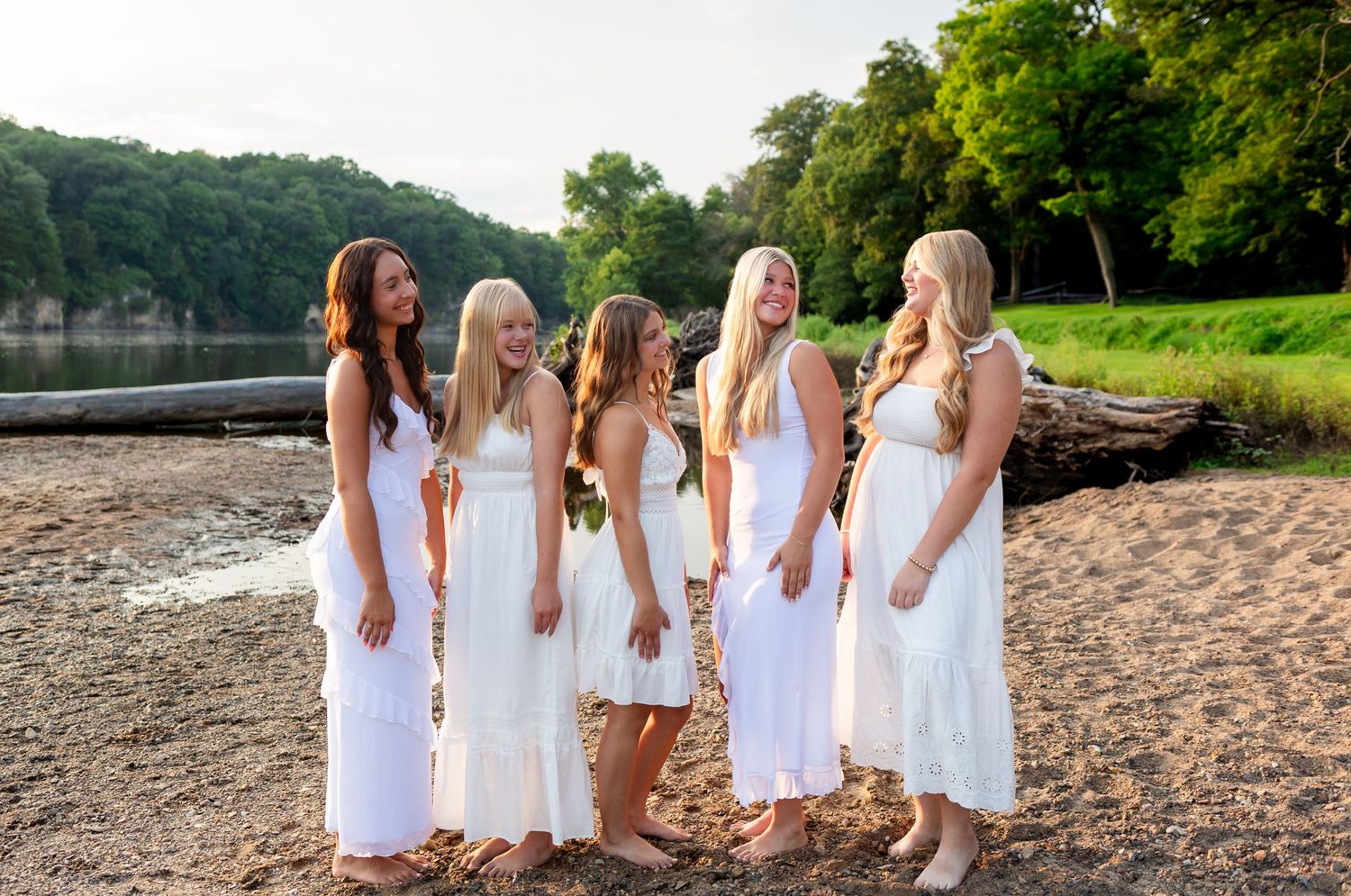Five high school senior girls in flowing white dresses stand on a sandy beach at Palisades-Kepeler park with trees in the background.