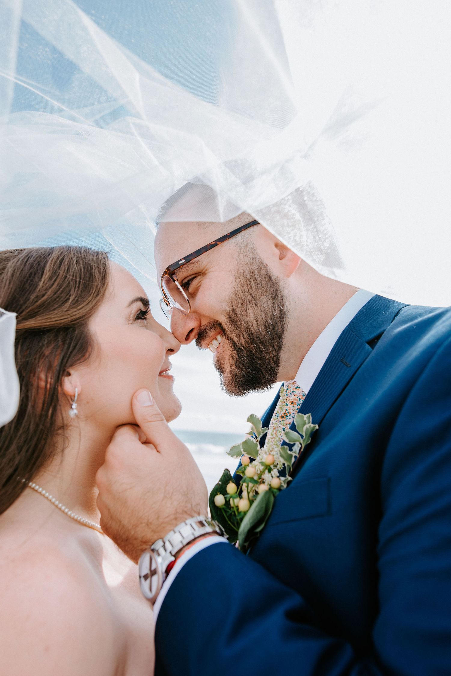 A romantic wedding moment captured under a flowing veil as a couple shares an intimate laugh together outdoors.