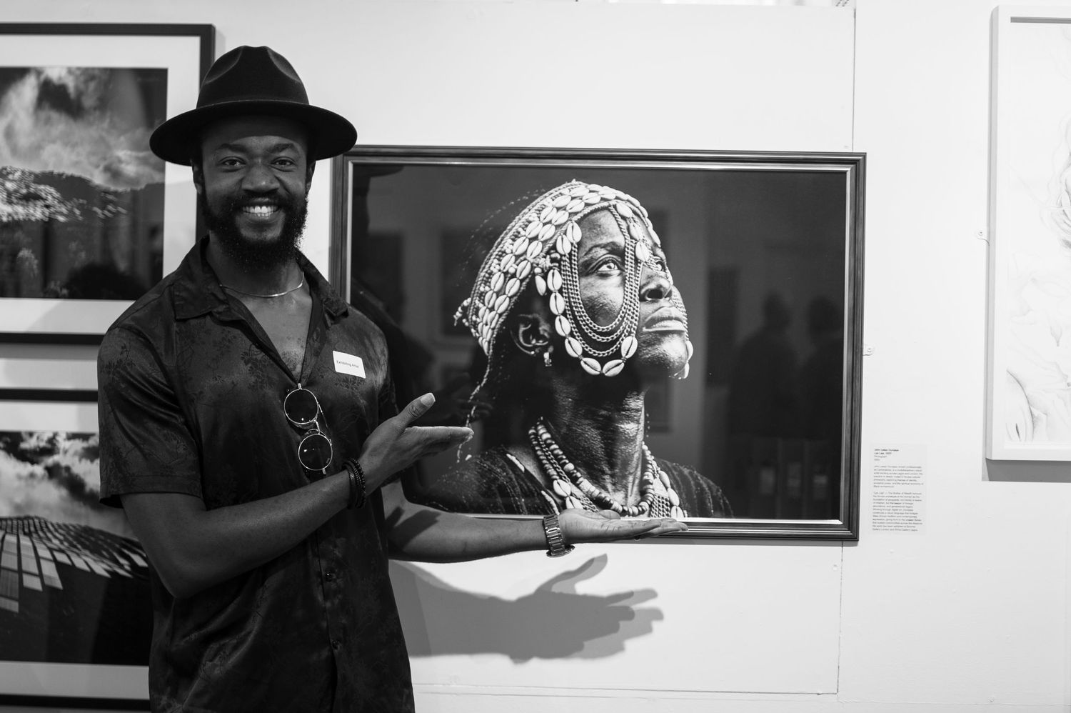 Man in black hat smiling beside a framed portrait photo of an African woman in traditional attire at an art gallery.