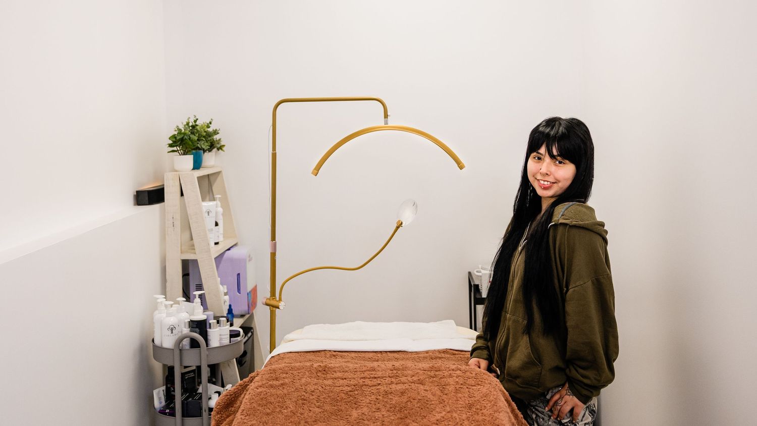 Woman with black hair smiles in a modern treatment room with a massage table and gold wall art.