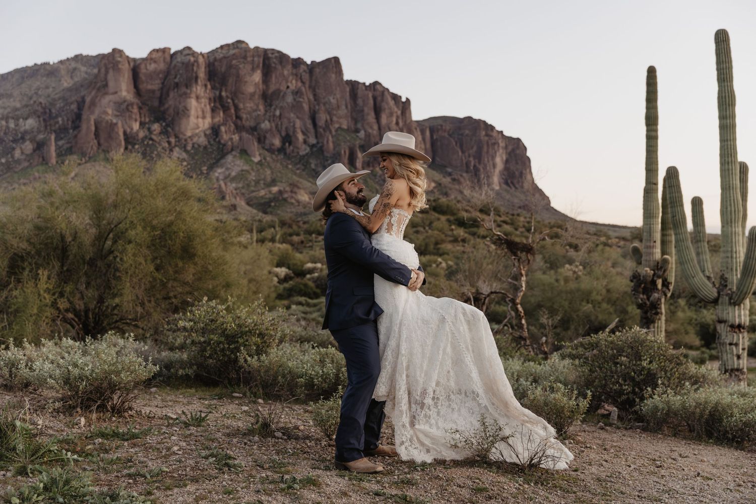 Bride and groom embrace in Arizona desert with saguaro cacti and Superstition Mountains at sunset.