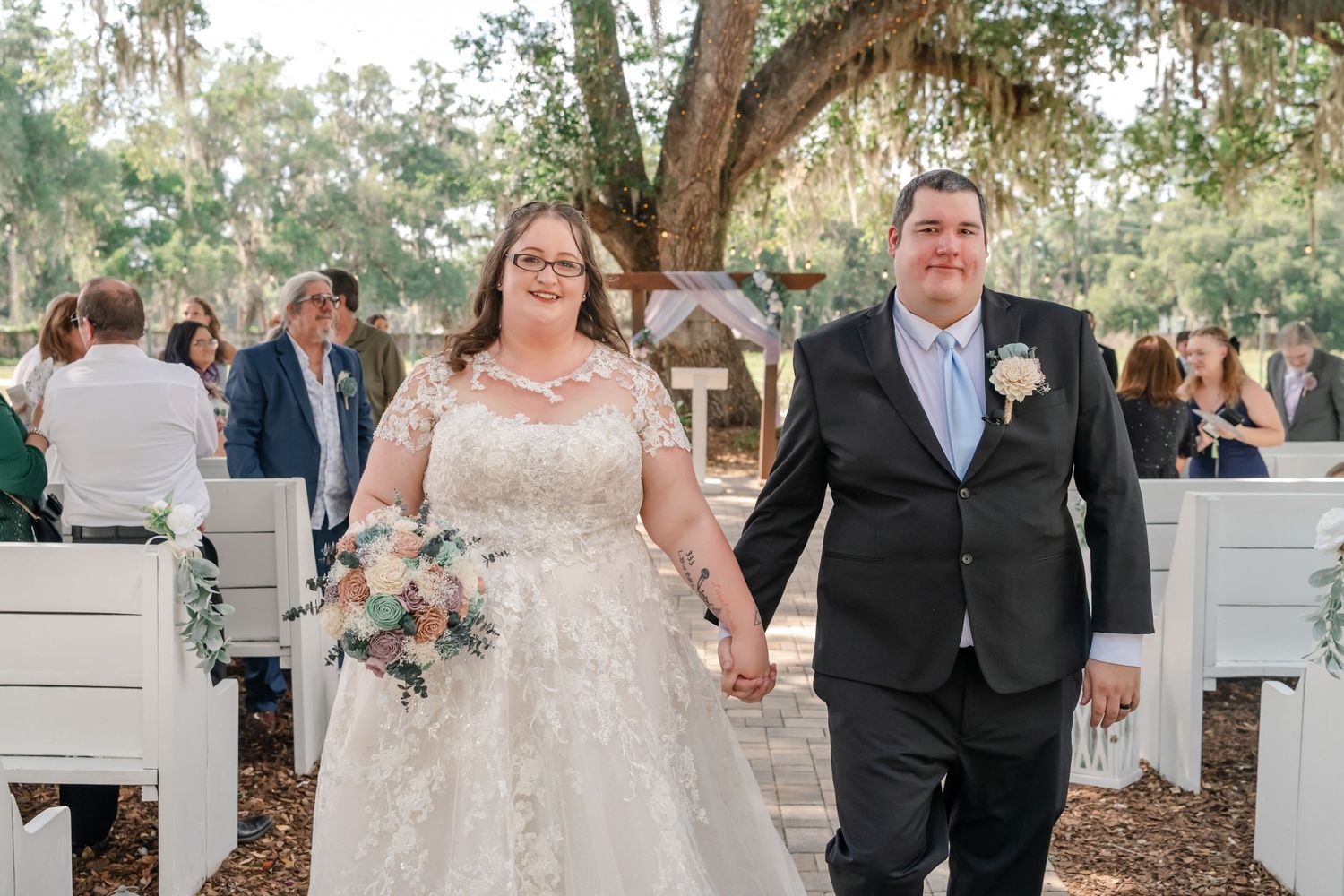 Newlywed couple holding hands walking down the aisle at an outdoor wedding ceremony under oak trees.